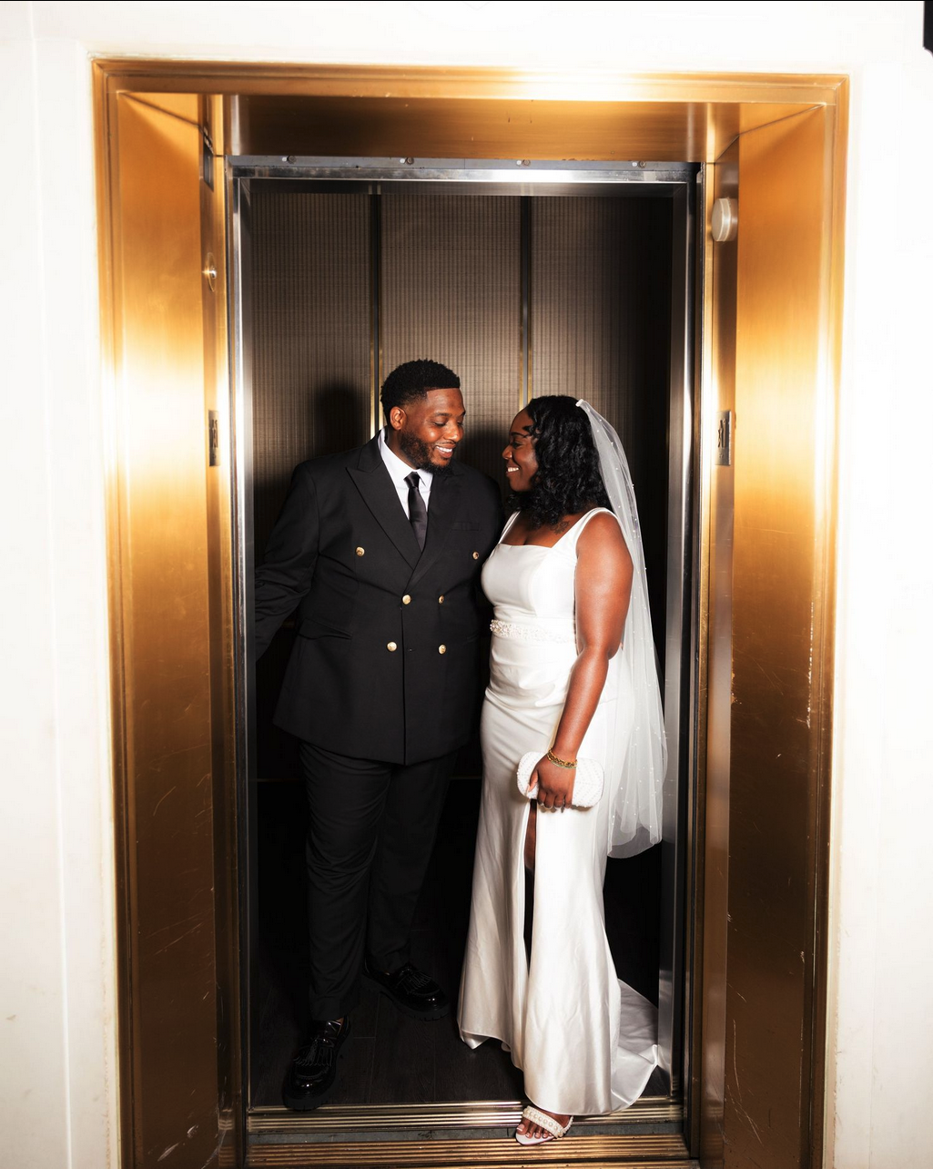 Bride and groom smiling at each other inside a gold elevator