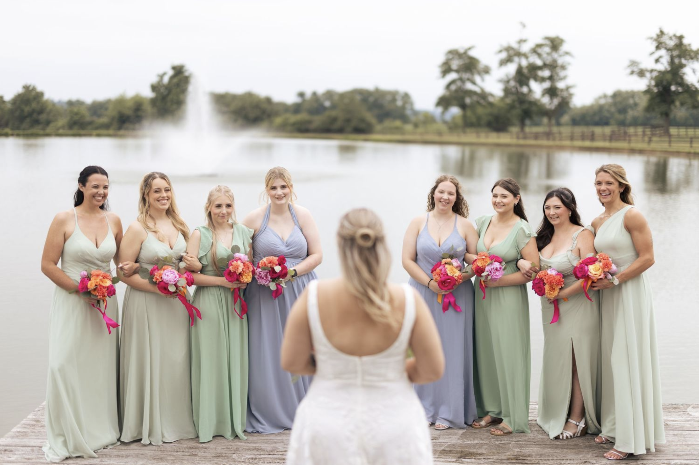 Bride facing her bridesmaids in green and blue dresses on a dock by a lake