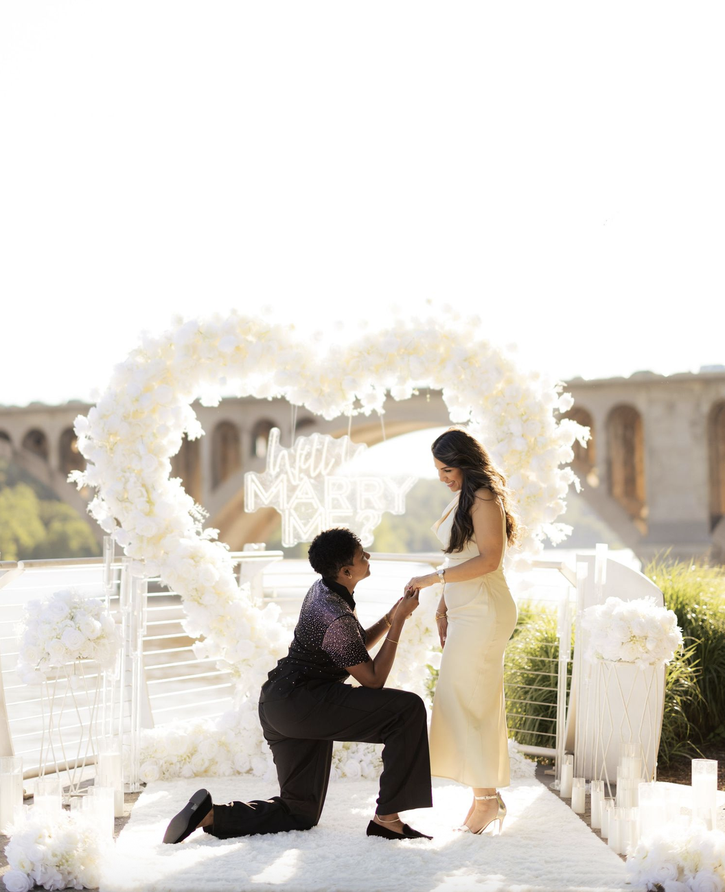 Proposal under a white heart-shaped floral arch with a bridge in the background