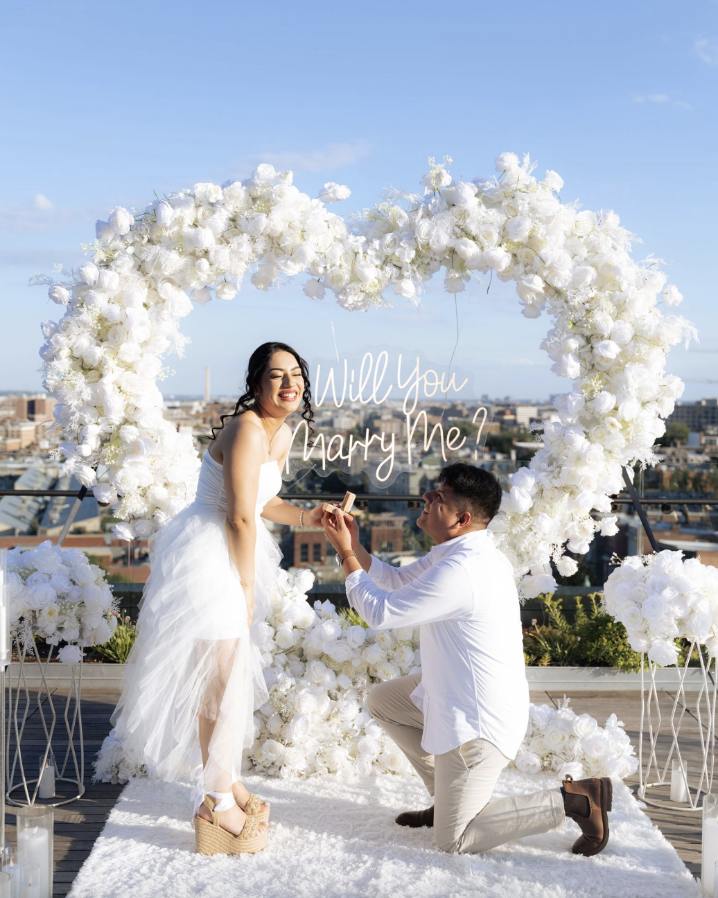 Man proposing under a heart-shaped white floral arch on a DC rooftop